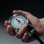 A hand holding a sphygmomanometer showing blood pressure readings in a medical setting.